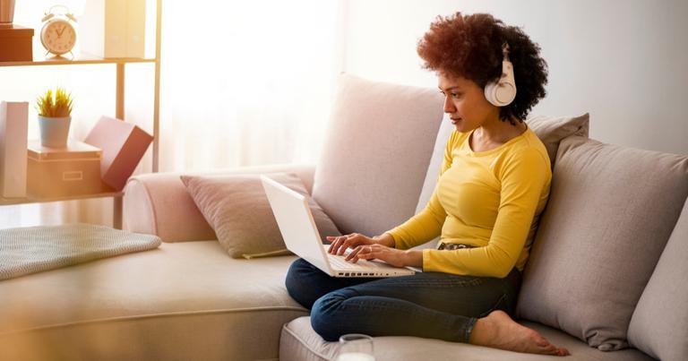 Mujer sentada en el sofá de su casa disfrutando de una partida de casino online con su ordenador portátil mientras escucha música por los auriculares.