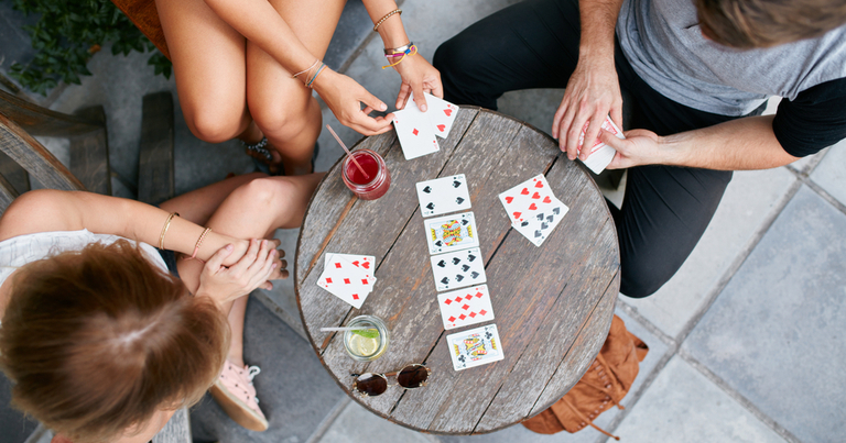Tres jóvenes adultos juegan a las cartas en una mesa de madera al aire libre.
