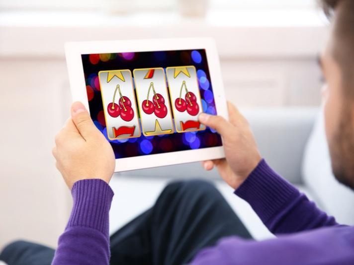 Hombre jugando a una tragaperras online en una tableta donde aparecen cerezas, números siete y estrellas.