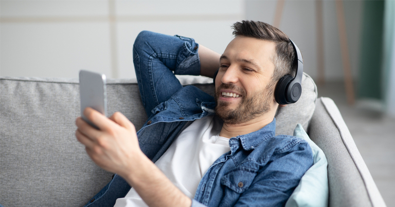 Un hombre feliz sentado en el sofá con su teléfono móvil mientras escucha música con auriculares.